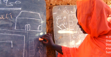 The photo shows a woman from Sudan standing infront of a blackboard with a chalk in her hand, writing something. She is wearing an orange dress that also covers her head.