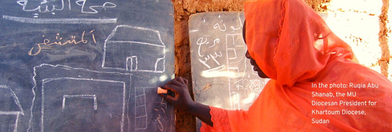 The photo shows a woman from Sudan standing infront of a blackboard with a chalk in her hand, writing something. She is wearing an orange dress that also covers her head.